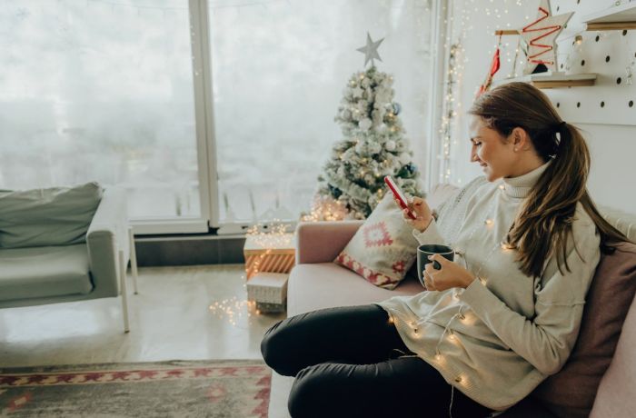 A woman curled up on the sofa looking at her phone with mug in hand christmas lights around her