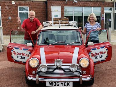 a man and a woman standing either side of a classic red mini cooper on a self drive road trip