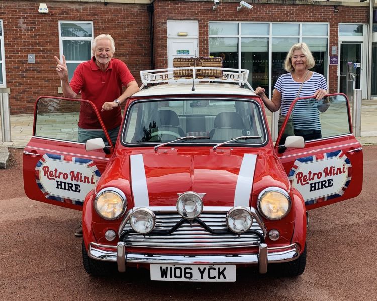 a man and a woman standing either side of a classic red mini cooper on a self drive road trip