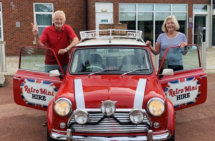 a man and a woman standing either side of a classic red mini cooper on a self drive road trip