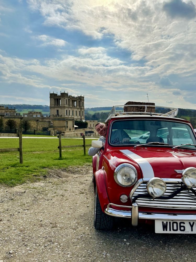 a man poking his head out of the window of a red classic mini cooper parked up outside chatsworth house