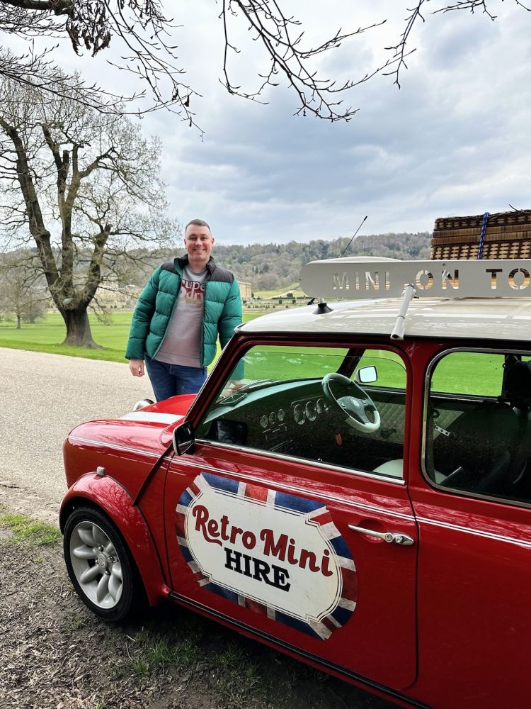 a man standing in front of a red classic mini cooper in the grounds of chatsworth house
