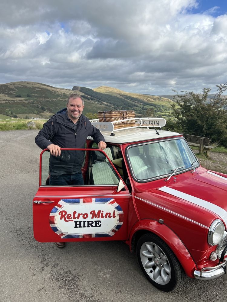 a man standing alongside a red classic mini cooper with rolling hills in the background