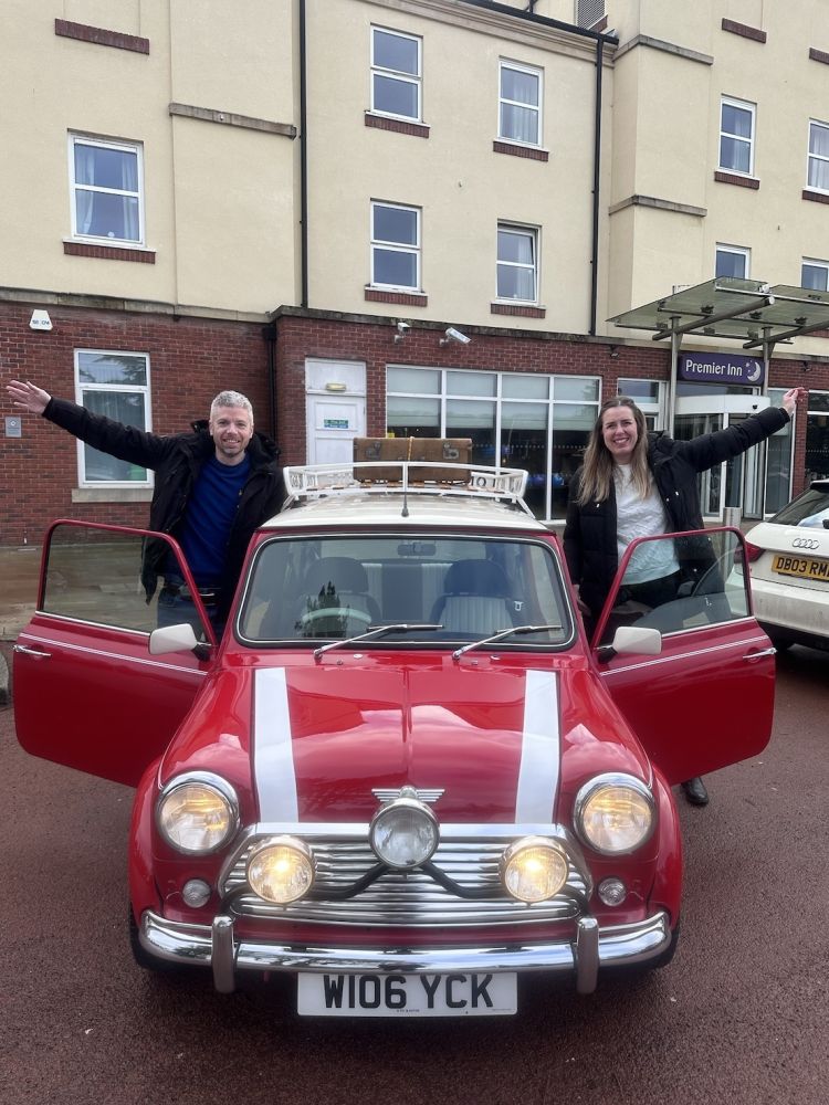 a man and a woman standing either side of a red classic mini cooper ready for a self drive road trip