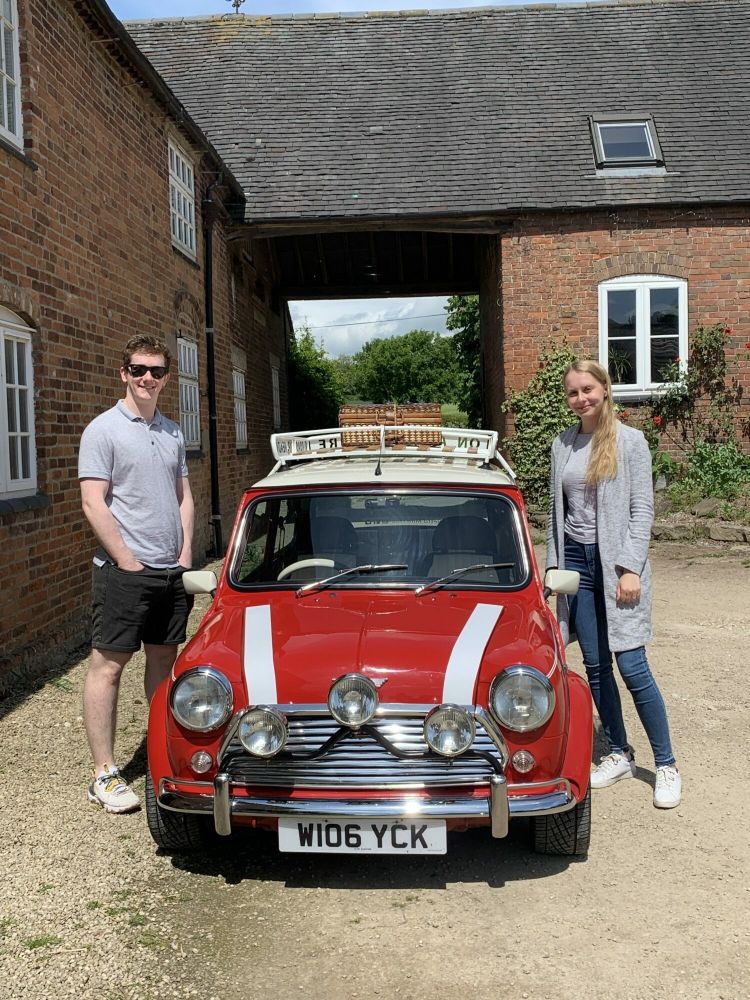 a man and a woman standing either side of a red classic mini cooper ready for a hire and drive experience