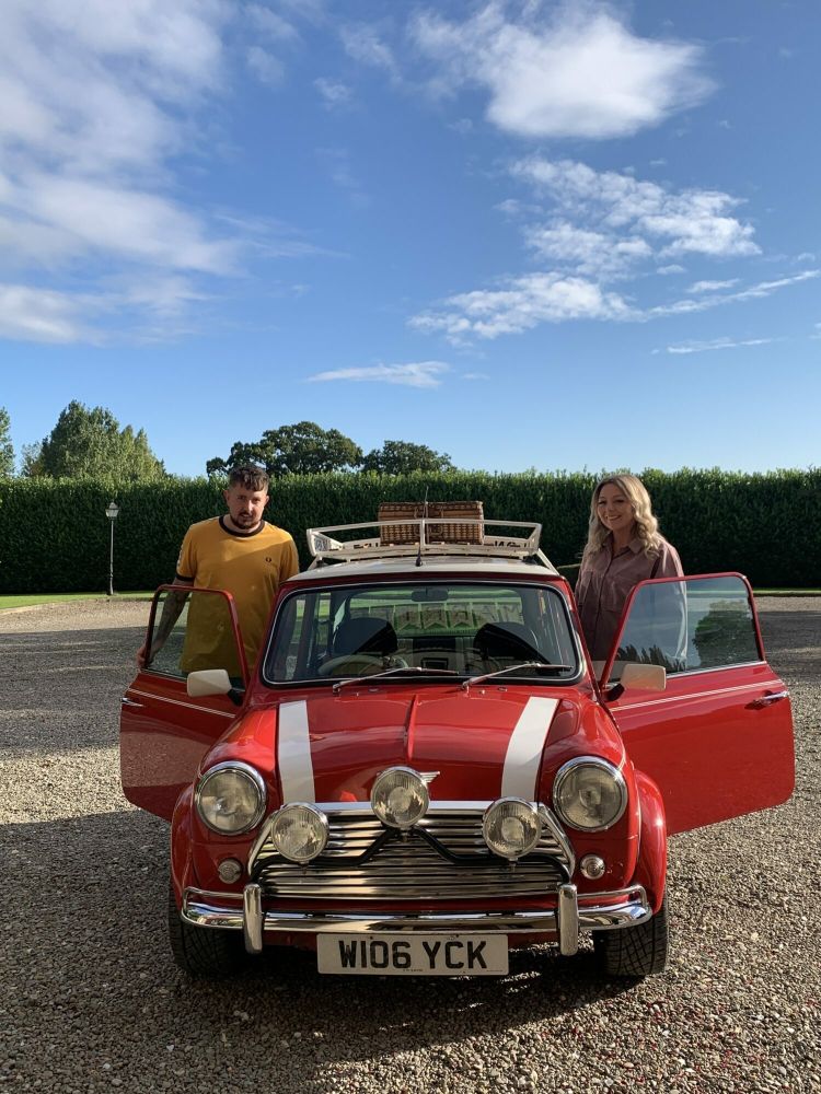 a man and a woman standing either side of a red classic mini cooper ready for a hire and drive experience