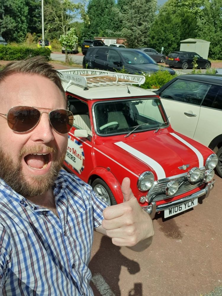 a man looking at the camera giving the thumbs up with a red classic mini cooper behind him