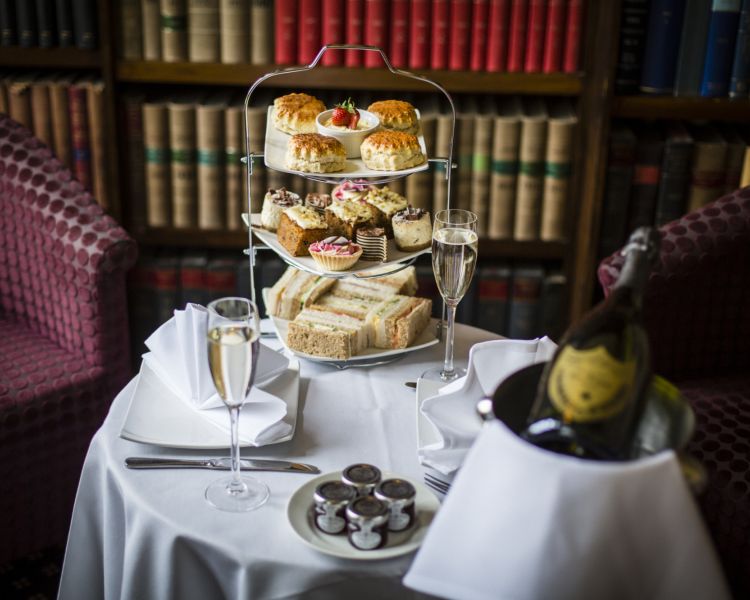 afternoon tea set up on a round table with white table cloth, a cake stand full of cakes and sandwiches and a bottle of champagne in an ice bucket