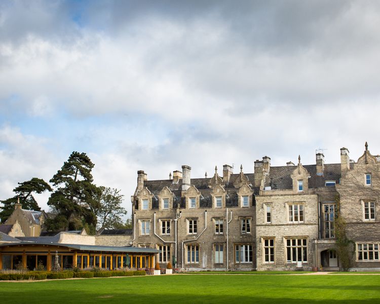 view of the back of a countryside manor house with a green lawn in the foreground leading up to the grey brick building