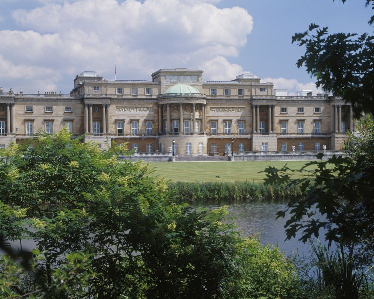 view of buckingham palace taken from the garden with trees and lake in the foreground then a lawn and then the palace