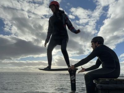 woman standing on a pump foil board with instructor sitting on the side of the water