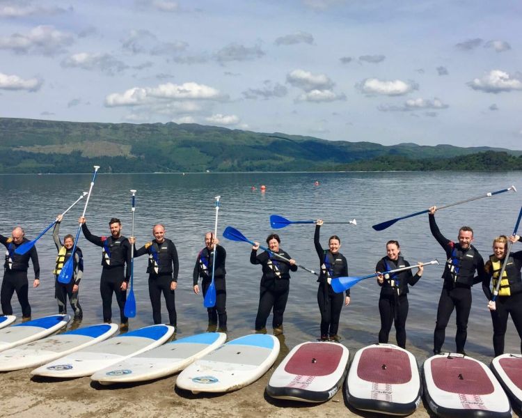 people lined up at the edge of a lake to go paddle boarding