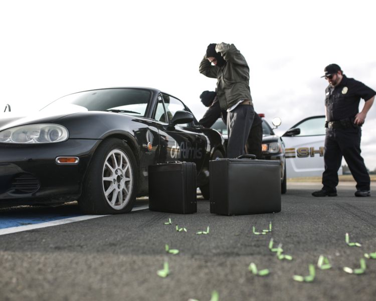 two people standing by the side of a black sports car with two briefcases on the floor with a policeman to the right of them