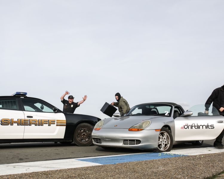 a police car with a man next to it holding his arms up next to a silver sports car with two people getting out, one is holding a briefcase