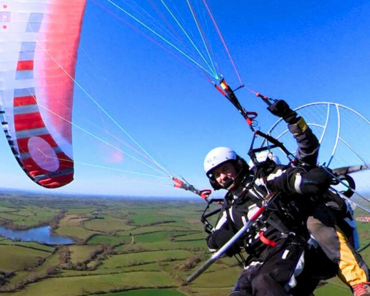 man on a tandem paramotor experience flying over green fields against a blue sky