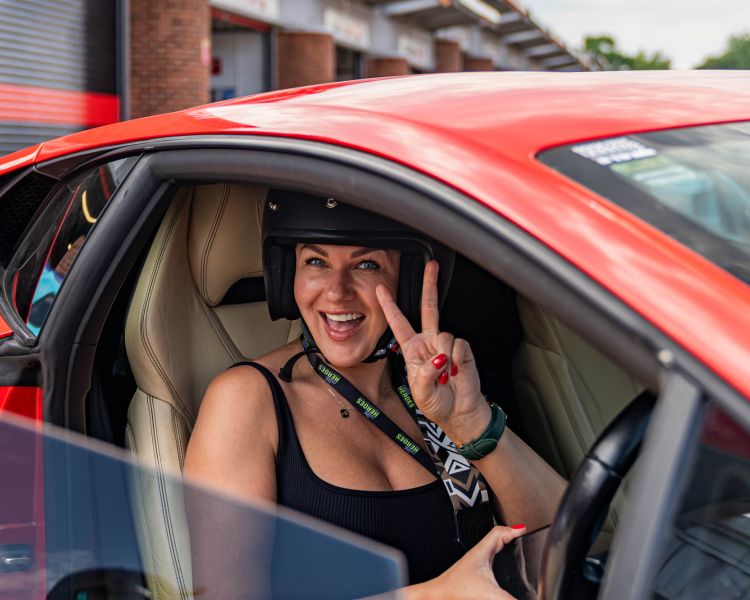 a woman smiling and giving the peace sign sitting in the drivers seat of a supercar on a driving experience day
