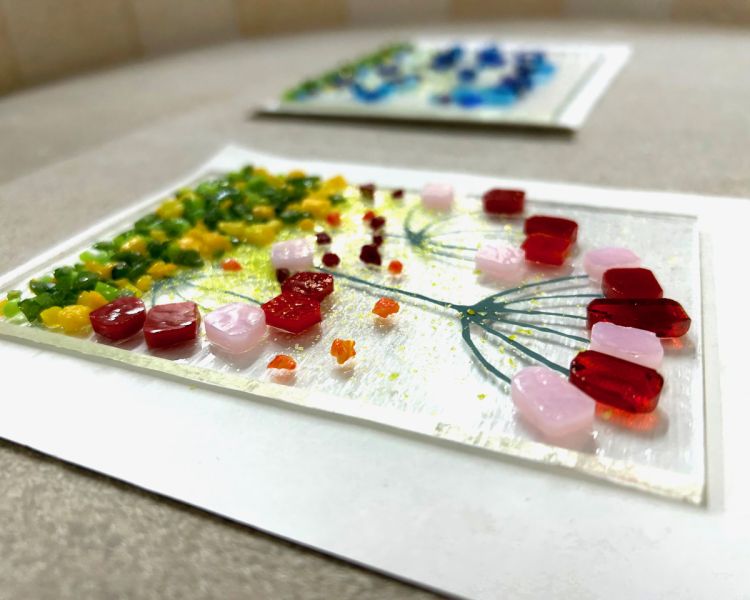 the front of a glass tea light holder with a coloured floral design laid on a board ready to go into the kiln