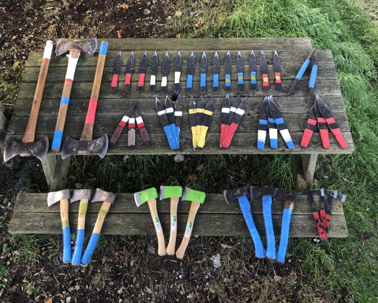 a selection of axes and blades with coloured handles laid out on a wooden table as part of an axe throwing experience