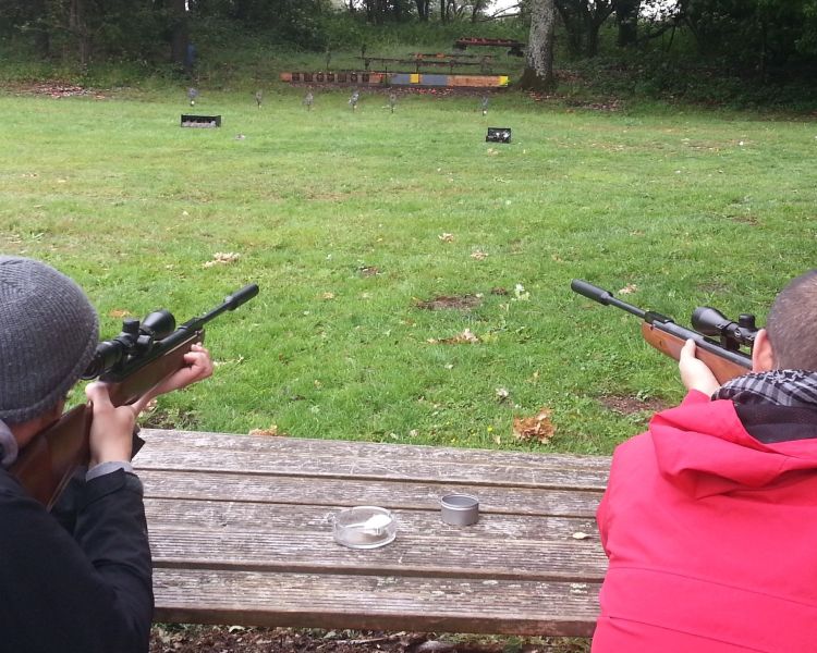 view from behind of two people sitting at a wooden table pointing air rifles at a target as part of am air rifle shooting experience
