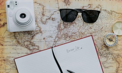 A map with a compass black sunglasses a white camera and a notebook open on top of it with bucket list written on the page