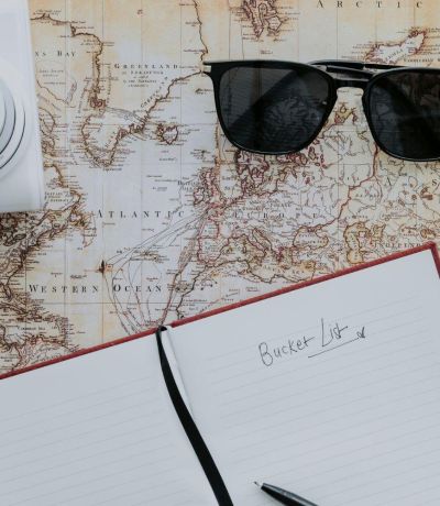 A map with a compass black sunglasses a white camera and a notebook open on top of it with bucket list written on the page