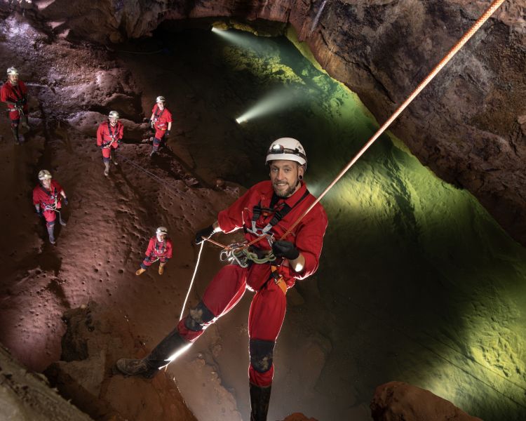 man abseiling into cave with others waiting at bottom