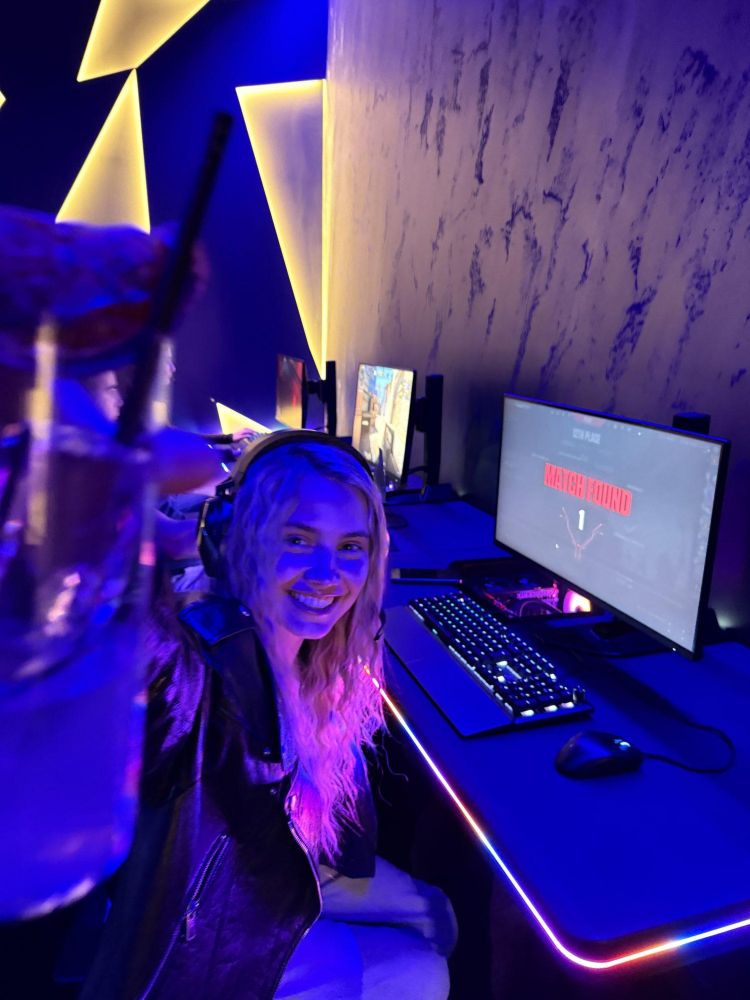 girl holding up cocktail sat at pc at gaming bar in london
