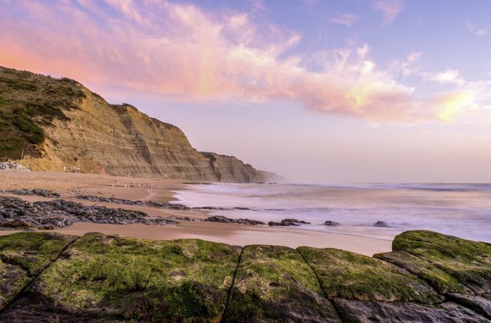 beach surrounded by the sea and cliffs covered in mosses under a cloudy sky during the sunset