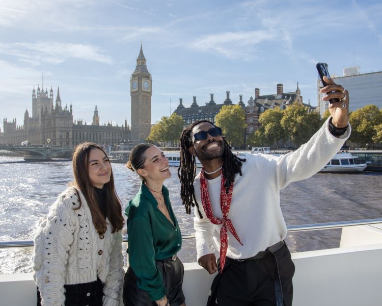 three people taking a selfie onboard a thames river cruise with big ben in background
